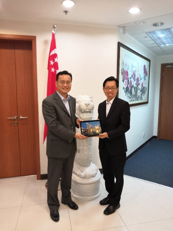 Two men in suits holding tablet with building image. Singapore flag and lion statue are visible.